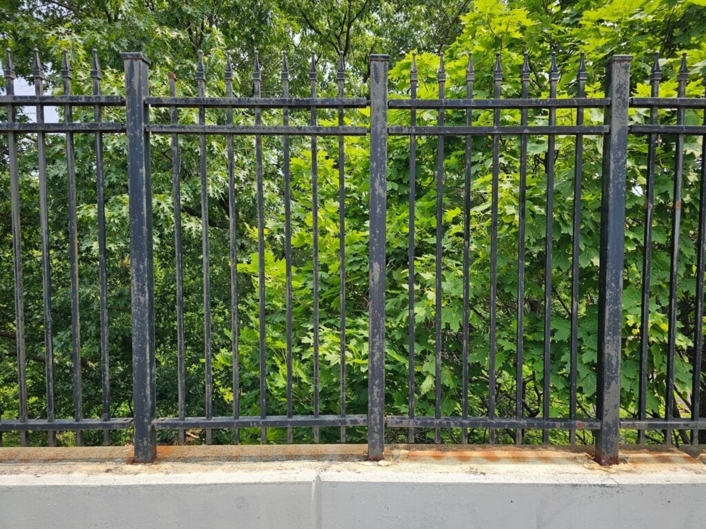 Today, we're looking at a relatively typical type of steel fence that set on top of a concrete substrate. This particular substrate happens to be in the form of a curb which raises the level of the fence slightly above the adjacent grade. Whether fences or partitions are made of masonry, concrete, steel, or wood, they all require upkeep and maintenance. Nowadays they even make some types of fences from PVC, fiberglass, or even synthetic materials which require less maintenance. All of these different types of materials require a degree of continuous upkeep and maintenance though and will all deteriorate on their own, naturally. They all need to be connected into the ground or on top of the hardscape or paving. Here, in mounting to the curb, the steel has been embedded into the concrete. As far as aesthetic decisions are concerned, there's almost no question, the classic iron and steel look is one of the best options for places like here in Washington DC where we are surrounded by historic and significant architecture. It allows for a bit of openness, but it looks substantial and more elegant than wood fences. Masonry walls can be built around perimeters of spaces, but masonry will usually completely block the area from visual openness. Masonry happens to look beautiful. It has a timeless classic quality. Often, that isolated and separated feel is needed, depending on the nature of the space. Particularly in backyards or courtyards where you want a feel of privacy, masonry is one of the best options. In contrast, if you want the space to feel open yet still have separation from access, steel and classic iron work is probably the best option. In this particular installation, the 2x2 steel posts have been embedded deep into the concrete substrate. There is a curb that sets the limit to the transition of the landscaping. These posts are set not just into the curb but even farther down beyond the depth of the curb itself. In this case the curb is only about 7.5 inches tall. With an embedment of only 7.5 inches into the concrete, it gives a post like this very little resistance from any type of contact and even low resistance from typical weather events like high winds. A deeper insertion will create structural resistance through a cantilevered embedment that can resist lateral forces. That type of structural resistance is often called moment resistance or rotational resistance. The words themselves may seem a little bit confusing. When you think of the words used here for this type of structural resistance, you might automatically think of things slightly differently, just from intuition. Starting with the concept of moment resistance, that's the type of resistance that is achieved by inserting these posts down into the concrete substrate. We say they are vertically cantilevered because although they are embedded into the concrete for purposes of structural resistance, they stick out and project beyond the concrete and that resistance extends with them. Interestingly, the resistance decreases relative to the distance by which they protrude and extend away from the point of structural mooring. When you refer to this as a moment resistance, in structural terms, you think of time. A moment is a word that describes a brief amount of time. Here though, although it sounds very different, it actually has a similar root, etymologically speaking, in the definition of the word. In the early centuries of the understanding of physics, people referred to the movement of objects with a root word that's shared with the word moment. The understanding of that type of force, related to movement, was based on motion which was also connected, not just to the change of position, but also to the rate of change, relative to time. We also think there's a little bit of confusion that people also experience when calling this a rotational force resistance. That type of rotational force resistance refers to the individual post's resistance to tilt over. It doesn't specifically refer to the posts resistance from twisting around rotationally. That difference is important because they resist movement or force very differently depending on the positioning of the particular object. The embedment and the interconnection between the adjacent rails and panels of the steel offers significant resistance to twisting. Only the post, though, offers resistance to the actual rotational force that poses risk to the fence. That type of rotational force is related to pressure applied to the vertical position of the plane of the fence. Since our company, Dupont Tuckpointing and Masonry, specializes in masonry restoration, historic brick repointing, and tuckpointing services in the Washington D.C. area, we make it a point to study these types of issues. These buildings are uniquely historic, and their preservation requires skilled masons who are technically trained in the best practices and knowledge of proper restoration techniques. We understand the significance of maintaining the architectural integrity of these historic structures, and our team of experienced professionals is dedicated to delivering exceptional craftsmanship. Whether you require masonry restoration, tuckpointing, or brick repointing services, we are here to help. At Dupont Tuckpointing and Masonry, we take pride in our work and strive to ensure that every project is executed with the utmost care and attention to detail. We are committed to preserving the rich heritage of Washington D.C.'s built environment for generations to come. If you have any questions or needs regarding masonry restoration, historic brick repointing, or tuckpointing services, please do not hesitate to reach out to us. We would be delighted to assist you and provide you with the expertise and quality workmanship that your historic property deserves. You can reach us by telephone at (202) 796-7644 and you can reach us by email from the contact form on our website at https://duponttuckpointingmasonrydc.com/contact-us/.
